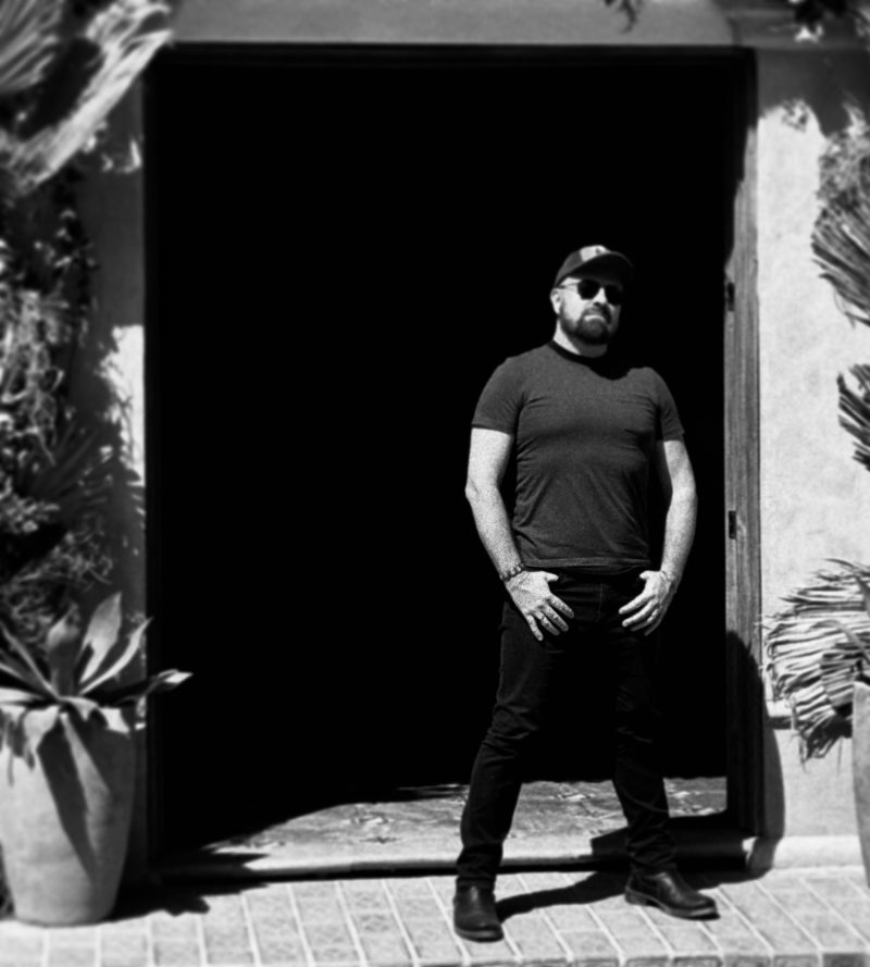 Séa Byrne Standing in front of a doorway in rulral, Jalisco Mexico. He's wearing a cowboy cap shades and boots.